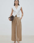 A woman poses against a plain background wearing the SUNCOO Gilem Vest layered over a sleeveless white V-neck top, high-waisted wide-leg beige trousers, white sandals, and holding a large black clutch for added elegance.