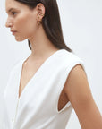 A woman with straight dark hair, wearing the SUNCOO Gilem Vest—a sleeveless white V-neck top with subtle shoulder pads—is shown in profile against a plain light background, accessorized with multiple gold hoop earrings.
