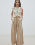 A woman stands against a plain background wearing SUNCOO's Woven Pantalon Jeff—high-waisted darted wide-leg pants with pleats—paired with a sleeveless white crochet top and crisp white shoes, her hands in her pockets.