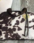 The Faire Oakley Overnighter- Faux Cowhide is a vegan travel bag featuring a brown and white cowhide pattern, dark handles, and an "alema" tag with ribbon, displayed before a light mannequin and rustic wooden background.