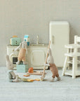 Three small Maileg plush bunnies play in a vintage-style kitchen, featuring the Maileg Bread box w. utensils, fridge, stove, sink, highchair, and dollhouse touches; two bunnies mop while another stands by the sink.