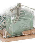 A Maileg Bread box w. utensils by Maileg is wrapped in clear plastic with a ribbon, displayed on a wooden tray next to a patterned card and a small green tin mailbox—charming accessories for any dollhouse.