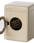 The Maileg Washing Machine by Maileg is a small, beige appliance with a round, left-opening front door that reveals a black perforated drum. It features three buttons and a rectangular panel on the top front—ideal for laundry day play.