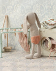 A Maileg fabric bunny doll in orange shorts stands by the Maileg Drying Rack with pegs and a toy washing machine, hanging tiny clothes amid folded fabrics and miniature laundry items against patterned wallpaper.