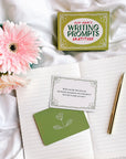 A gold pen lies on an open notebook beside a pink flower and Rupi Kaur's Writing Prompts Gratitude cards by Simon & Schuster Inc. One card asks about a recent act of kindness and its impact.