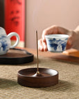 A lit incense stick rests in the Faire Walnut Wood Incense Burner on a woven mat, while a hand holds a blue-and-white teacup. Handcrafted ceramic plates with rustic glaze complete this serene, artisan-inspired tea scene.
