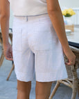A person wearing a white shirt and Frank & Eileen's Waterford Short with a wide-leg fit stands indoors by a wooden table and wicker chairs, their back to the camera.