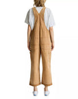A person with shoulder-length, wavy hair stands with their back to the camera, wearing Denimist’s RELAXED OVERALL in tan—a 100% cotton style featuring adjustable buckle straps, large back pockets, and a relaxed-leg silhouette—layered over a light gray t-shirt and paired with white sneakers.