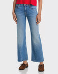 The lower half of a person stands against a white background, wearing Mother Lil Bookie Twist Skimp mid rise flare-leg jeans and brown moccasin-style shoes with a red top.
