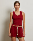 A woman with light brown hair tied back stands against a plain wall, wearing the Perfectwhitetee Layla Terry Short and a sleeveless red tank top. The shorts feature french terry cotton, an elastic waistband, and a white drawstring. She looks neutral.