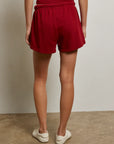 A person stands indoors facing away, wearing a sleeveless red top and Perfectwhitetee's Layla Terry Short with an elastic waistband, paired with white slip-on shoes on a concrete floor against a plain light wall.