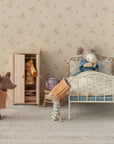 Three Maileg Mum Suit mice relax in a vintage-style bedroom by Maileg: one sits in bed sipping from a mug, one stands near a wardrobe with tiny outfits, and another wears pajamas and a hat. A teapot rests on the bedside table.