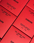 Several red rectangular boxes labeled "SUNBURN 12 oz candle" by Faire are arranged diagonally, showcasing a bold, non-toxic, vegan candle collection.
