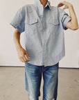 The individual is dressed in The Western Shirt from Hey Gang, an oversized light blue short-sleeved denim shirt with a classic vintage aesthetic, featuring two chest pockets. They pair it with blue jeans and have their arm raised slightly against a plain background.