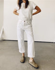 A person stands with hands on hips, wearing a white t-shirt, Hey Gang's "The Loose Crop" jeans made of 100% cotton denim, and brown slip-on shoes. The scene features a plain white wall and concrete floor, with the person's face out of view.