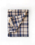 The Faire Cook's Cloth, a beige and navy blue plaid apron with cream straps, made from a cotton-linen blend, is shown folded on a white background.