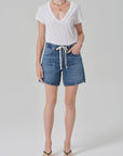 A person wearing Citizens of Humanity/AGOLDE brynn drawstring short in avant (lightwt vintage), a white V-neck T-shirt, flip-flops, a white belt, and layered necklaces stands against a plain light background.