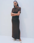 A woman with curly hair stands against a plain background, arms crossed, wearing White + Warren's Luxe Cotton Skirt in dark brown with a side slit and a matching Italian knit top. She completes the look with light pointed shoes.