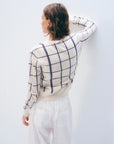 A person with short brown hair faces a white wall, wearing the White + Warren Cashmere Windowpane Cardigan and straight-fit white pants with large back pockets. One arm is bent, touching the wall.