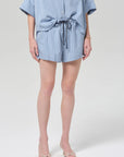A person wears the Citizens Of Humanity/AGOLDE este drawstring short in capri stripe—blue, silky-smooth shorts with a light blue button-up shirt and white open-toe sandals, standing against a light gray background.