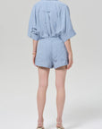 A person with light skin and short hair wears Citizens Of Humanity/AGOLDE's este drawstring short in capri stripe, styled with a matching vertical striped light blue shirt and white low-heel sandals, standing against a plain light gray background.