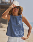 A woman smiles on a sunny beach, wearing a light sun hat, dark sunglasses, and the Frank & Eileen Shell Tank with white shorts. She holds her hat brim as the ocean shimmers in the background.