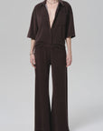 A person models the Citizens Of Humanity/AGOLDE nora pull on pant—a loose, dark brown, wide-leg style—paired with a matching short-sleeve button-up shirt, standing barefoot against a plain light gray background.