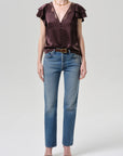 A person wears a short-sleeve, deep v-neck dark satin blouse with lace trim, Citizens Of Humanity Savannah Cigarette Jean in Patina, black heeled sandals, and a gold necklace, standing against a plain light gray background.