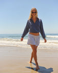 A woman wearing sunglasses, a blue long-sleeve top, and Faire’s Stripe Track Short walks barefoot along the sunny shoreline, with waves and a clear blue sky in the background.