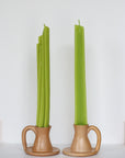 Two light brown ceramic holders cradle Faire’s handmade Celery Beeswax Candles—one green taper is bent and uneven, the other stands straight—displayed against a plain white background.