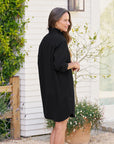 A woman with long brown hair stands outside, smiling sideways by a potted plant and white house in lush greenery, wearing the Frank & Eileen Mary Classic Shirtdress-Ruffle with black long sleeves.