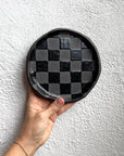 Black Check Snack Plate by Faire with a hand holding it against a textured white wall.