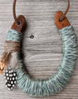 The Lucky Horseshoe by Faire features an upcycled horseshoe wrapped in blue-grey twine, brown string, and feathers, hanging from a leather cord—a unique housewarming or personalized gift.