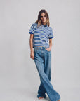 A woman stands against a plain background wearing Denimist's SHRUNKEN POLO, a blue and white striped cotton t-shirt, paired with loose-fitting blue jeans. Her left hand rests in her pocket while her right arm is relaxed at her side.
