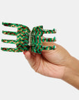 A hand holds the Faire Ursa Claw in Andi, a green and orange patterned acetate hair accessory, against a plain white background.