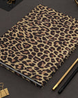 The Faire Leopard Journal with a hardcover design rests on a textured dark surface beside gold and black pencils, gold binder clips, and geometric desk decor.