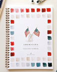 The Faire Watercolor Workbook, a spiral-bound beginner guide titled "Americana," features two small American flags in a glass jar on the cover with red, blue, and gray watercolor swatches, shown on a table with paint supplies.