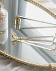 A Faire Candle Snuffer rests with two gold wick trimmers on an ornate oval mirror tray near a sunlit window, showcasing elegant and functional candle accessories for stylish home decor.