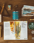 A flat lay of vintage items on wood, featuring the Faire Saguaro National Park Candle, a slingshot, matches, blue can, postcards, flashlight, oil-finish wax, coins, and an open book with cactus florals and a Southwest US map.