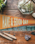 The Faire "Have a Good Day" Felt Pennant rests on a wooden surface amid plants, stones, and wood—this cheerful, motivational banner is perfect for brightening any space.