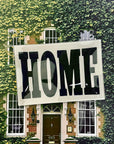 The Faire Home Block Decoupage Glass Tray, featuring vintage "HOME" typography, is displayed in front of a green ivy-clad house with brick walls, large windows, and a dark brown door.