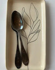 Two hand-etched silver spoons rest on Faire’s Floral Small Tray, a rectangular cream-colored piece featuring a minimalist black line drawing of a leafy branch. The spoons’ handles align perfectly with the base of the drawn branch.