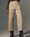 A person wears the Denimist Georgia Work Pant—beige, high-waisted, wide-leg pants with front patch pockets—with a brown long-sleeve top and black ankle boots, standing against a plain gray background.