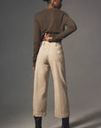 A person stands against a plain gray background, wearing Denimist's Georgia Work Pant—a high-waisted, wide-leg style—paired with a brown sweater and black ankle boots, with one hand on their hip.