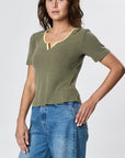 A woman poses in the Faire Stardust Hemp Top, a short-sleeved olive green shirt with a yellow-trimmed V-neck, paired with blue jeans, radiating retro charm against a plain light gray backdrop.