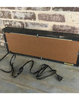 The Faire Lighted Sign features a rectangular design with corkboard back, black frame, and attached cord with switch and plug. Shown on a wooden surface against a brick wall.