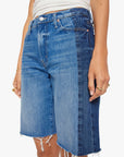A person wears the Mother Mid Rise Maven Bermuda Fray shorts featuring a light blue front, dark blue sides, raw frayed hems, metal button and pocket details, paired with a white ribbed tank top for a stylish wide leg look.