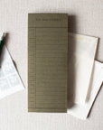 The Faire To Do Today Planner, a green daily notepad with hourly slots, is shown with a pen, postage stamps, and two envelopes on a light beige fabric surface.