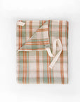 The Faire Cook's Cloth is a folded apron in a soft cotton-linen blend with green, beige, and orange plaid stripes and cream waist ties, shown on a white background.