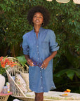 A woman smiles outdoors in the Frank & Eileen Mary Classic Shirtdress- Ruffle, featuring a flattering fit and ruffle collar. She holds sunglasses, surrounded by greenery, a white wicker chair, flowers, and a table with drinks and fruit.